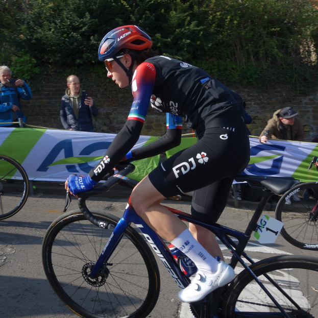 Eine Frau, identifiziert als Katie Archibald, trägt einen Helm, Schutzbrille, Handschuhe und Schuhe und fährt ein Fahrrad auf einer Straße neben einer Menge, mit einem Banner, einer Wand, Bäumen und einem Gebäude im Hintergrund.