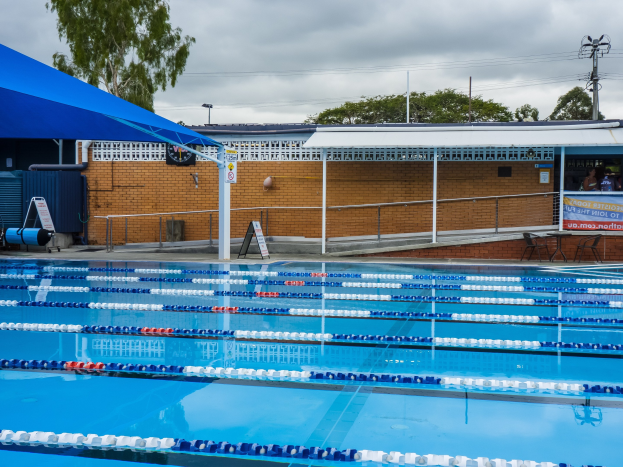 Großer Schwimmbad mit Bahnentrennern, umgeben von einem Zaun, einer Hütte, einem Banner, Stühlen, einem Tisch, einem Schild, Pfosten, einer Straßenlaterne, Drähten, Bäumen und einem bewölkten Himmel.