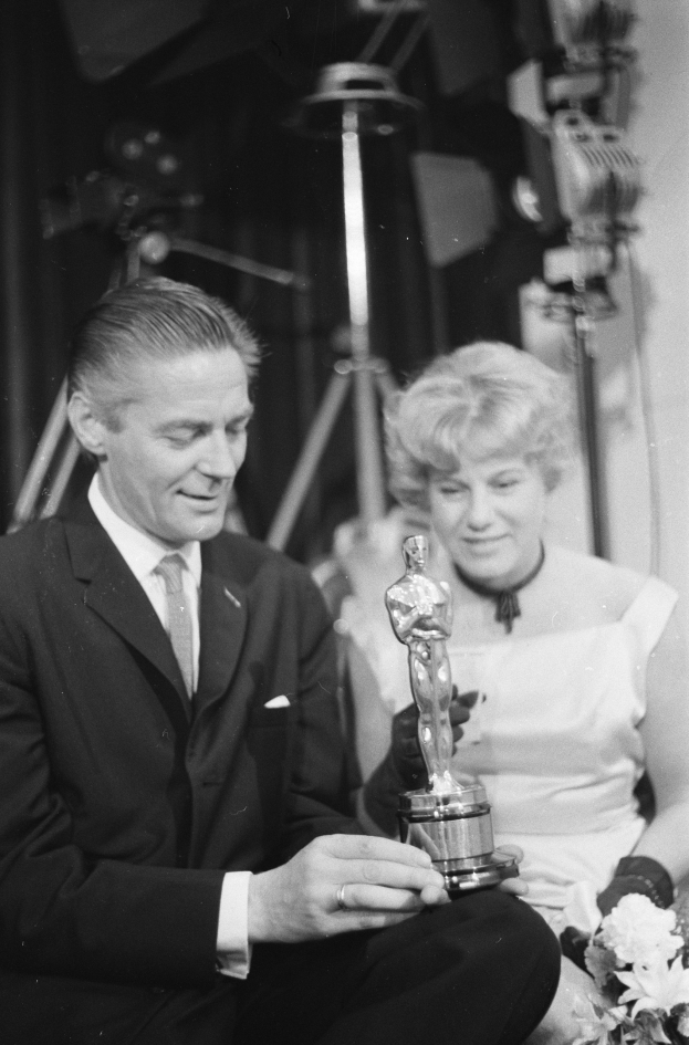 Ein Mann in einem schwarzen Blazer, weißem Hemd und Krawatte hält eine Oscar-Statue in der Hand und sitzt neben einer Frau, die einen Blumenstrauß hält, beide in Abendgarderobe, mit Lichtern im Hintergrund, dargestellt in Schwarz-Weiß.