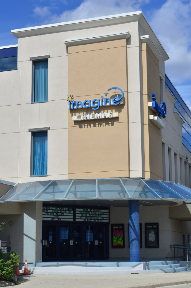 Außenansicht des Imagine Cinemas-Gebäudes in St. Louis, Missouri, mit Glasfenstern und -türen, einer Schautafel, einem Metallzaun, Pflanzen, einem Verkehrspfahl, Bäumen und einem bewölkten Himmel.