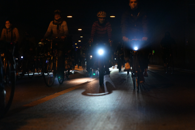 Eine Gruppe von Menschen, die nachts mit Fahrrädern durch eine Stadtstraße fahren, tragen Helme und werden von Stadtlichtern beleuchtet.