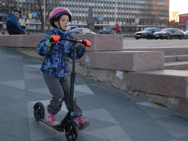 Ein junger Junge in Helm und Handschuhen fährt auf einem Roller eine Treppe hinunter, mit Stufen, Fahrzeugen, Menschen, Bäumen, Pfählen, Brettern, Gebäuden und einem klaren blauen Himmel im Hintergrund.