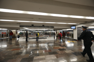 Eine Gruppe von Menschen, die durch eine belebte U-Bahn-Station geht, mit einem Polizisten im Vordergrund, umgeben von Geländern, Bildschirmen und Schildern.