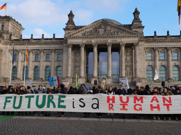 Eine Gruppe von Menschen hält ein Transparent mit der Aufschrift 'Zukunft ist ein Menschenrecht' vor dem Reichstaggebäude in Berlin, Deutschland, mit sichtbaren Fenstern, Säulen und Bögen des Gebäudes, umgeben von Fahnenmasten und einem bewölkten Himmel.