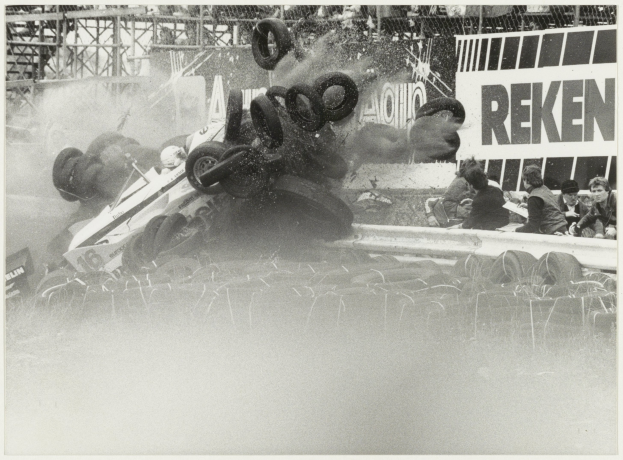 Schwarz-weiß-Foto eines verunglückten Rennwagens umgeben von Reifen, mit einer Gruppe von Menschen rechts und einer Tafel mit Text im Hintergrund.