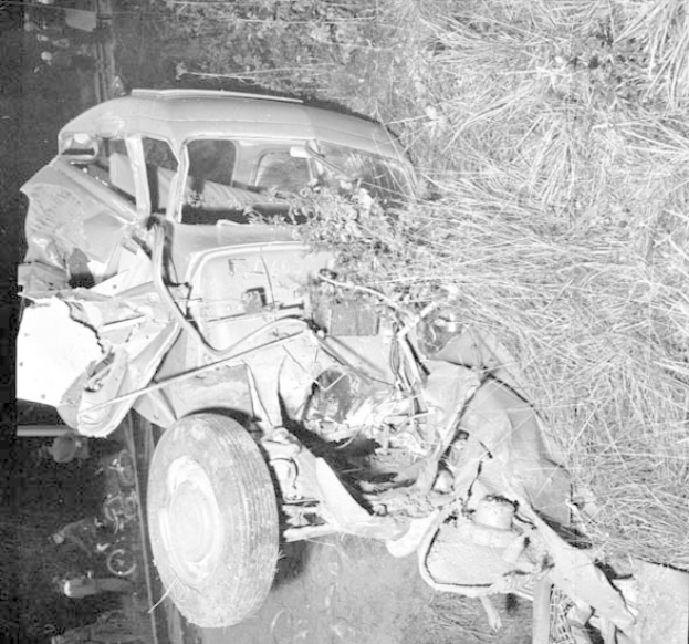 Schwarzes und weißes Foto eines schwer beschädigten Autos mit eingedrückter Front und zersplitterten Fenstern am Straßenrand, mit einem Baum und verstreuten Gegenständen im Hintergrund.