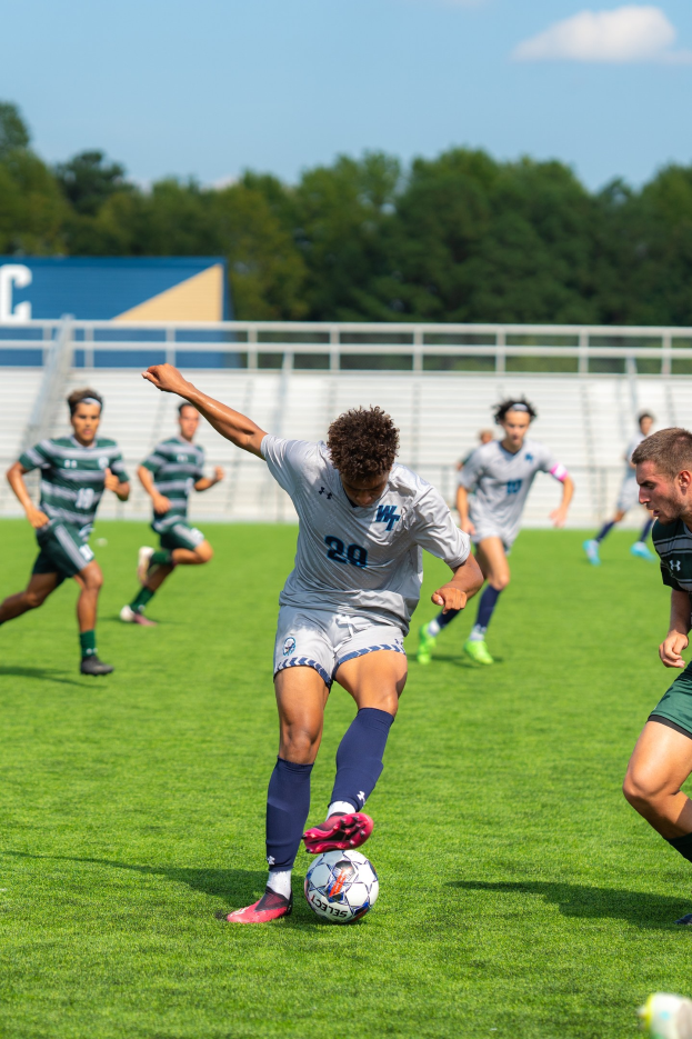 Junge Männer beim Fußballspielen auf einem Feld mit Bäumen und einem klaren Himmel, ein Spieler in Turnschuhen aktiv beteiligt.
