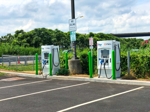 Elektrofahrzeug-Ladestation auf einem Parkplatz mit umgebender Infrastruktur, Grünfläche und einer Brücke im Hintergrund bei bewölktem Himmel.