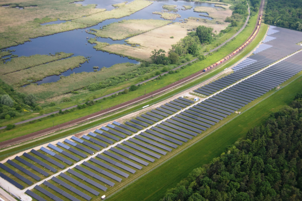 Luftaufnahme einer Solar-Farm mit Panels, umgeben von Bäumen, Gras, Pflanzen und Wasser, mit einem Zug auf einer nahen Bahnstrecke.