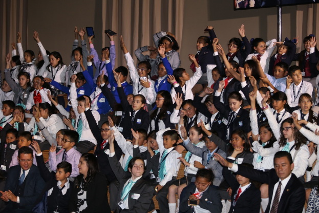 Gruppe von Kindern auf einer Bühne mit Vorhängen und einer Wanddekoration, während ein Mann und eine Frau vor ihnen stehen; ein Fernseher auf einem Ständer ist im Hintergrund sichtbar.