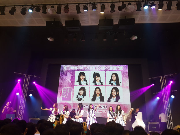 Eine Gruppe von Menschen, die auf einer hell erleuchteten Bühne performen, mit einem großen Bildschirm im Hintergrund, der Bilder von BK48 anzeigt, während eine Menge mit Kameras darunter zusieht.