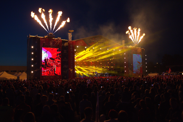 Eine große Menschenmenge auf einem Musikfestival schaut auf der hell erleuchteten Bühne Performern zu, mit Zelten links und Feuerwerk am dunklen Himmel dahinter.