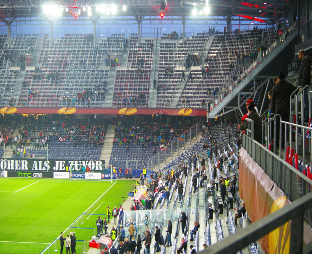 Großes Stadion voller Zuschauer bei einem Fußballspiel, mit sitzenden und stehenden Personen, einem Geländer, einer rechten Banner und einem leeren Hintergrund mit Deckenbeleuchtung.