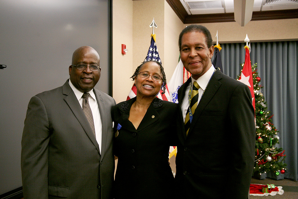 Eine Gruppe strahlender Menschen vor einem geschmückten Weihnachtsbaum und einem Vorhang, mit Fahnen und verschiedenen Gegenständen im Hintergrund und einem Tuch mit Gegenständen auf der rechten Seite.