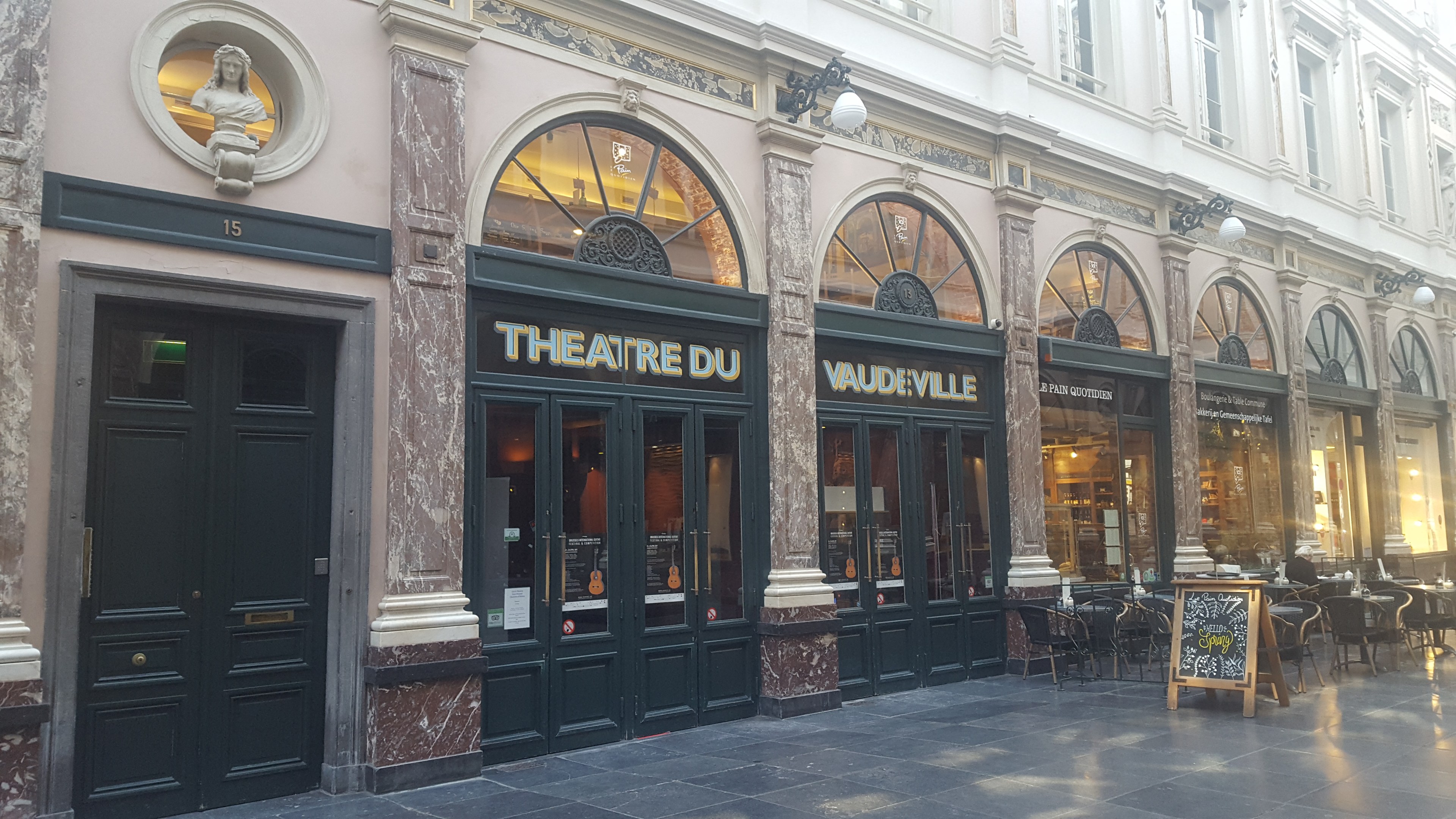 Das Bild zeigt die Außenseite des Théâtre du Vaudeville in Paris mit seinem Eingang, Fenstern, Beleuchtung, Beschilderung, Außenbestuhlung und einer Wandstatue.