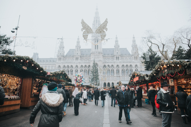 Ein lebendiger Weihnachtsmarkt in Wien, Österreich, mit Menschen, die herumlaufen, Ständen, die mit Lichtern und festlichen Décor geschmückt sind, einem Gebäude mit Fenstern im Hintergrund, Bäumen und einem klaren blauen Himmel.