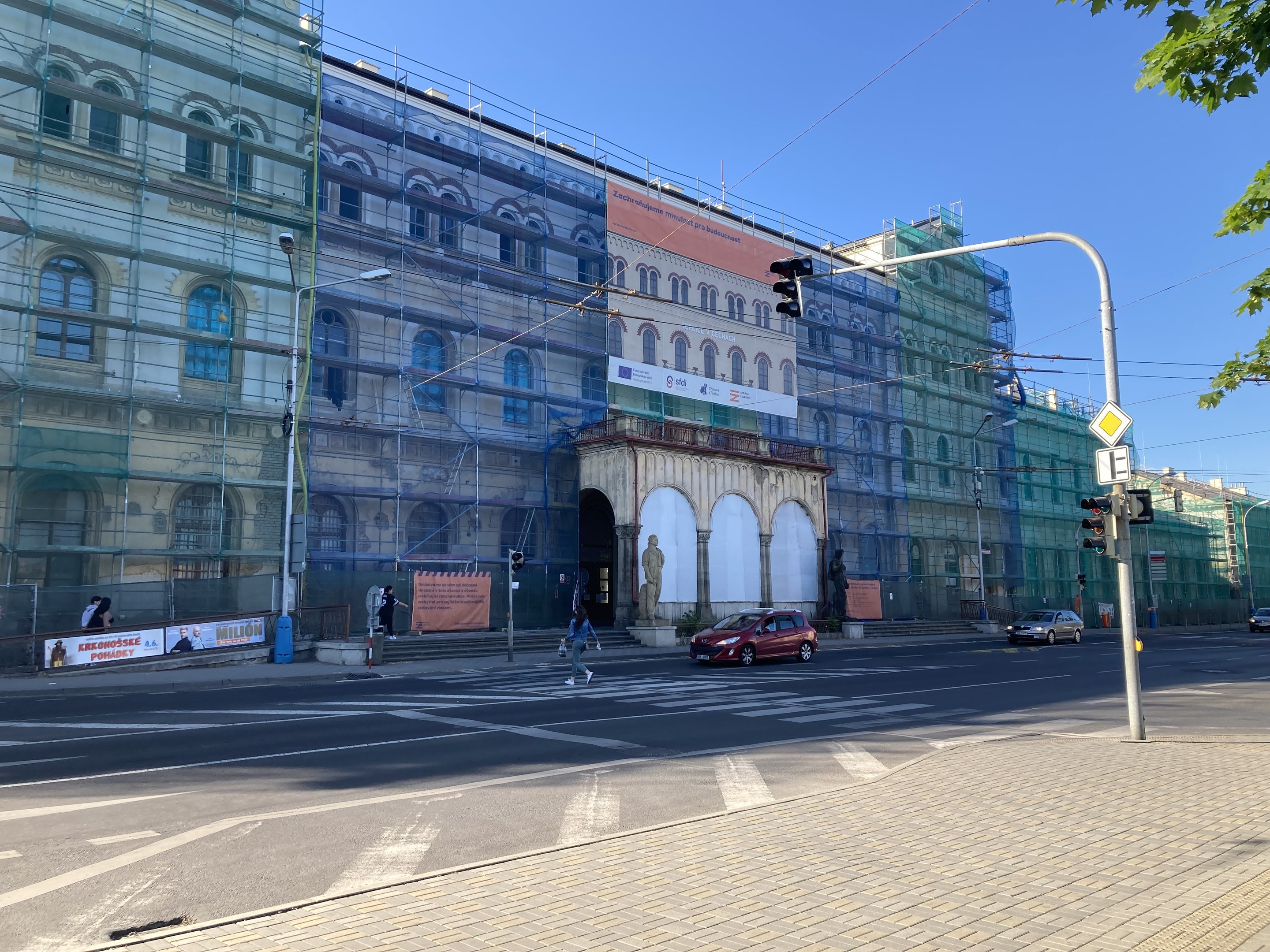 Großes Gebäude mit Gerüst während der Renovierung umgeben von Fahrzeugen, Fußgängern, Ampeln, Straßenschildern, Pfosten, Bäumen und einem klaren blauen Himmel.