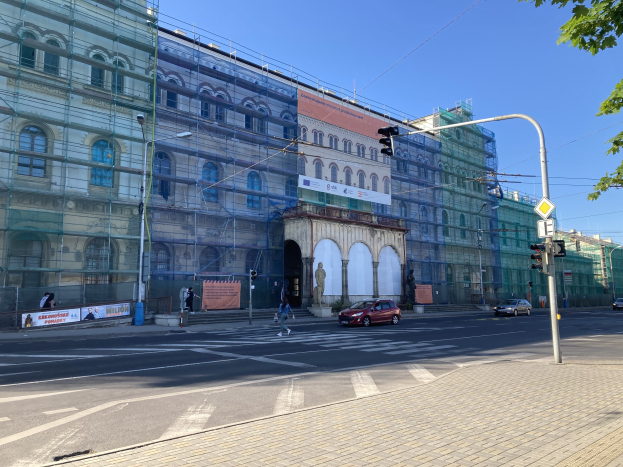 Großes Gebäude mit Gerüst während der Renovierung umgeben von Fahrzeugen, Fußgängern, Ampeln, Straßenschildern, Pfosten, Bäumen und einem klaren blauen Himmel.