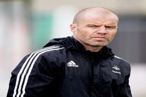 Ein kahlkopfiger Trainer in einer blauen Jacke mit weißen Streifen und einem weißen Kragen steht auf einem Fussballfeld und schaut ernsthaft nach vorne, die Hände verschränkt.