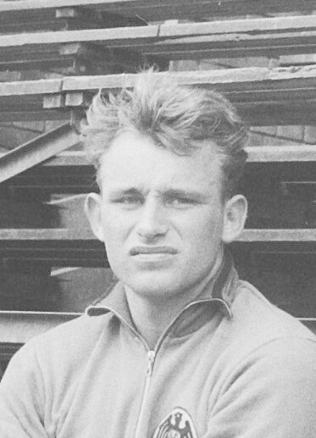 Ein Schwarz-Weiß-Foto eines Mannes in einer Jacke, der vor einem Stapel Holzplanken steht, der als legendärer deutscher Fussballer gilt.