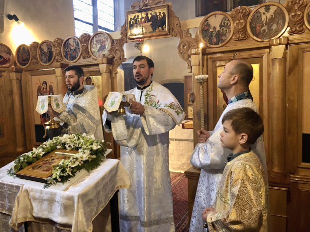 Gruppe von Menschen, die um einen Tisch in einer Kirche stehen, Bücher halten, mit einem Tuch und einem Blumenstrauß auf dem Tisch, Foto Rahmen und ein hölzernes Objekt an der Wand und ein Fenster im Hintergrund.