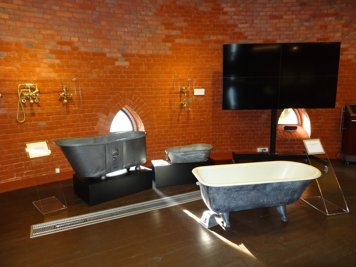 Ein kleines Badezimmer mit einer Ziegelwand, einer zentralen Badewanne, an der Wand montierten Wasserhähnen auf der linken Seite, einem an der Wand montierten Fernseher auf der rechten Seite und umgeben von Gegenständen, die eine gemütliche Atmosphäre schaffen.