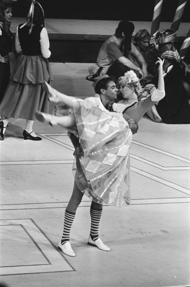 Ein Schwarz-Weiß-Foto eines Paares, das auf einer Bühne tanzt, mit ein paar Menschen auf der Treppe im Hintergrund und einem Pfahl im Vordergrund.