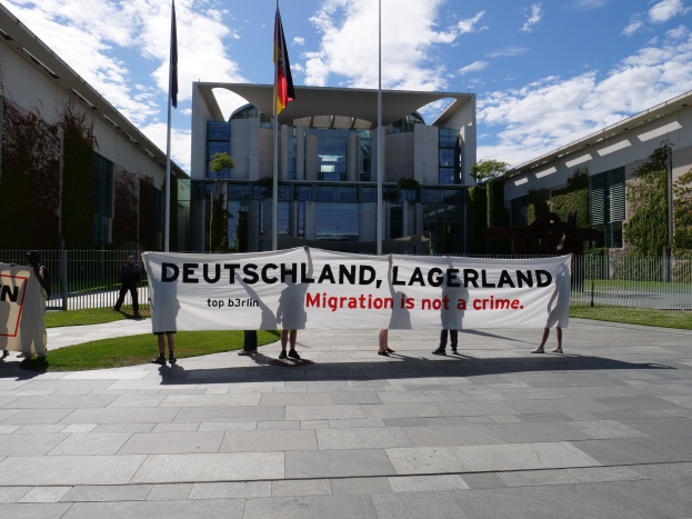 Gruppe von Menschen mit einem Banner "Deutschland, Lagerland Migration ist kein Verbrechen" vor einem Gebäude mit Fenstern, umgeben von Gras, einem Metallzaun, Pflanzen, Bäumen und Fahnenmasten.