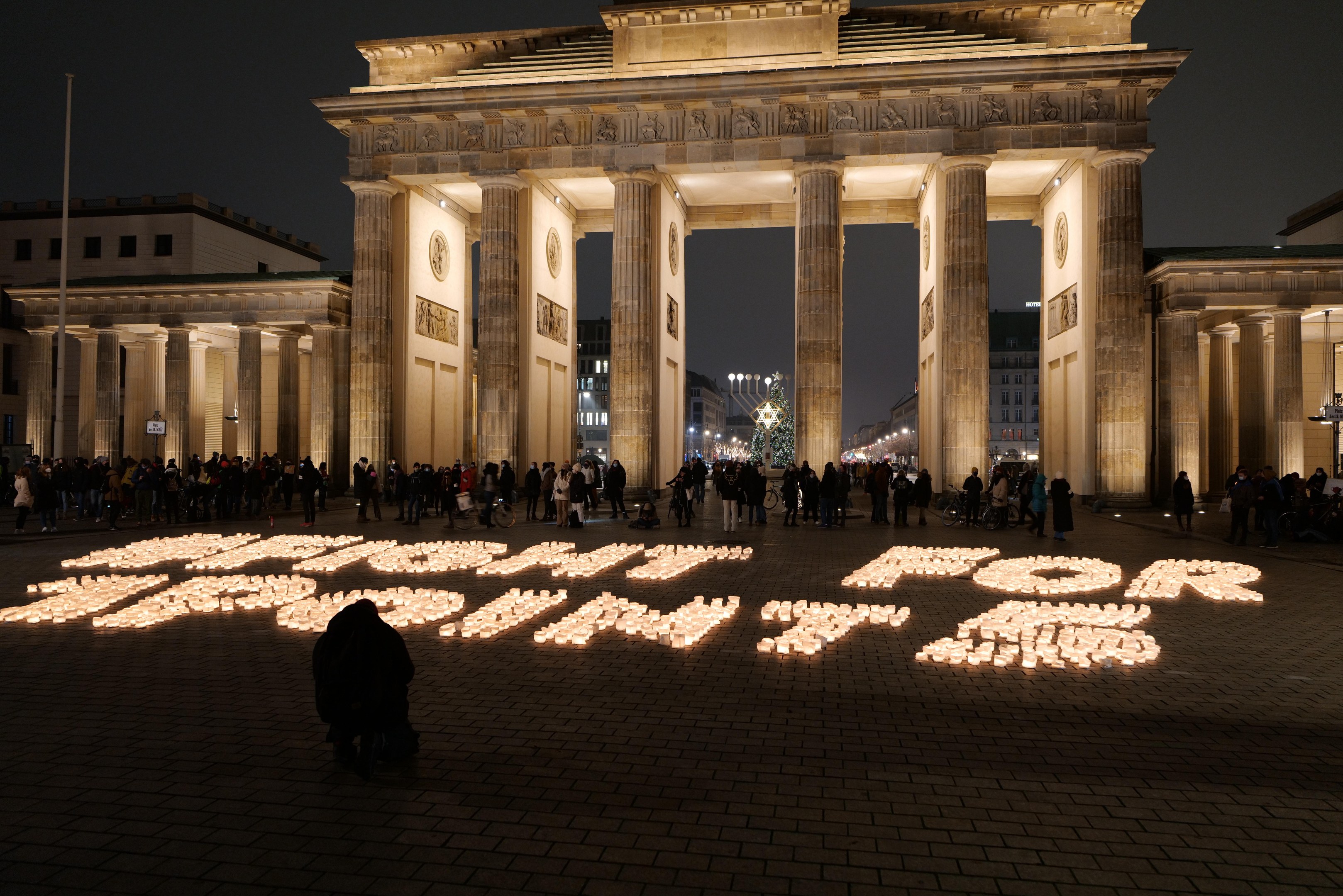 Eine Gruppe von Menschen steht vor dem beleuchteten Reichstag in Berlin, Deutschland, umgeben von Geb├Ąuden, Masten und Lichtern, und die Worte 'K├Ąmpfe für die Freiheit' sind auf den Boden geschrieben.