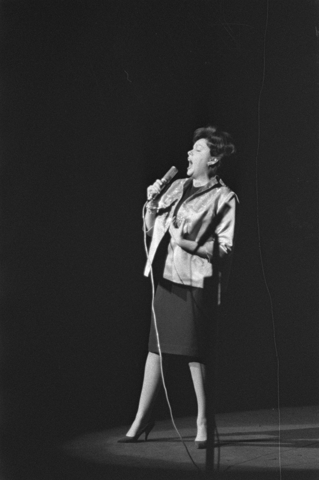 Eine Frau in einem schwarzen und weißen Outfit aus den 1950er-Jahren singt voller Freude auf einer von einem Scheinwerfer beleuchteten Bühne und hält ein Mikrofon.