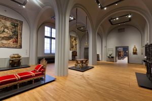 Ein geräumiger Raum im Kunsthistorischen Museum in Wien, Österreich, gefüllt mit Möbeln, paintings an den Wänden und dekorativen Elementen wie einem Sofa mit Kissen, Tischen mit verschiedenen Gegenständen, Säulen, Fenstern und Deckenleuchten.