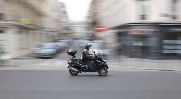 Eine Person in schwarzer Kleidung und einem Helm fährt auf einem Motorroller auf einer städtischen Straße mit anderen Fahrzeugen, Gebäuden und einem klaren blauen Himmel im Hintergrund.