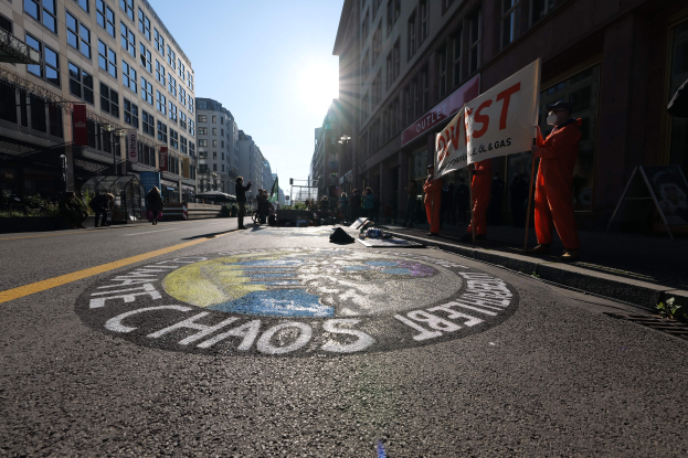 Eine Gruppe von Menschen mit Protestschildern und Plakaten auf einer Stadtstraße mit Gebäuden, Pflanzen und einer "Chaos"-Straßenmalerei, die auf Occupy Wall Street verweist.