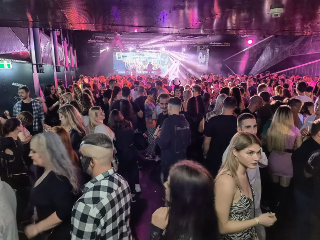 Eine große Gruppe von Menschen in einer Diskothek mit Deckenbeleuchtung und Säulen, mit einem Bildschirm im Hintergrund, der auf ein Absolut Nightlife-Event hinweist.