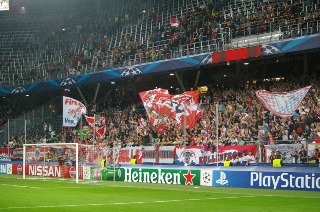 Ein Fußballspiel in einem hell erleuchteten Stadion mit einer großen Zuschauermenge, einem grünen Rasen, einem Tor, Bannern und einem Bildschirm in der oberen linken Ecke.