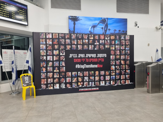 Eine große Wand zeigt mehrere Fotos, einen gelben Stuhl, israelische Regierungsflaggen auf Stangen, eine Tafel mit dem Text "Bring Them Home Now", einen montierten Bildschirm, Deckenleuchten und andere Gegenstände.