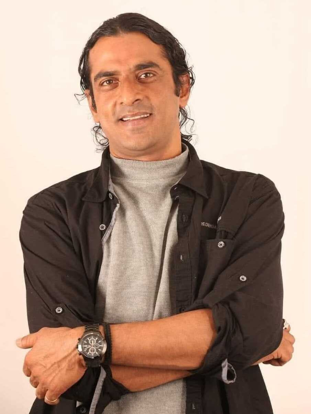 Ein Mann in einem schwarzen und aschfarbenen Kleid mit verschr├Ąckten Armen, der eine Uhr tr├Ągt und gegen einen wei├čen Hintergrund steht, der nachdenklich wirkt.