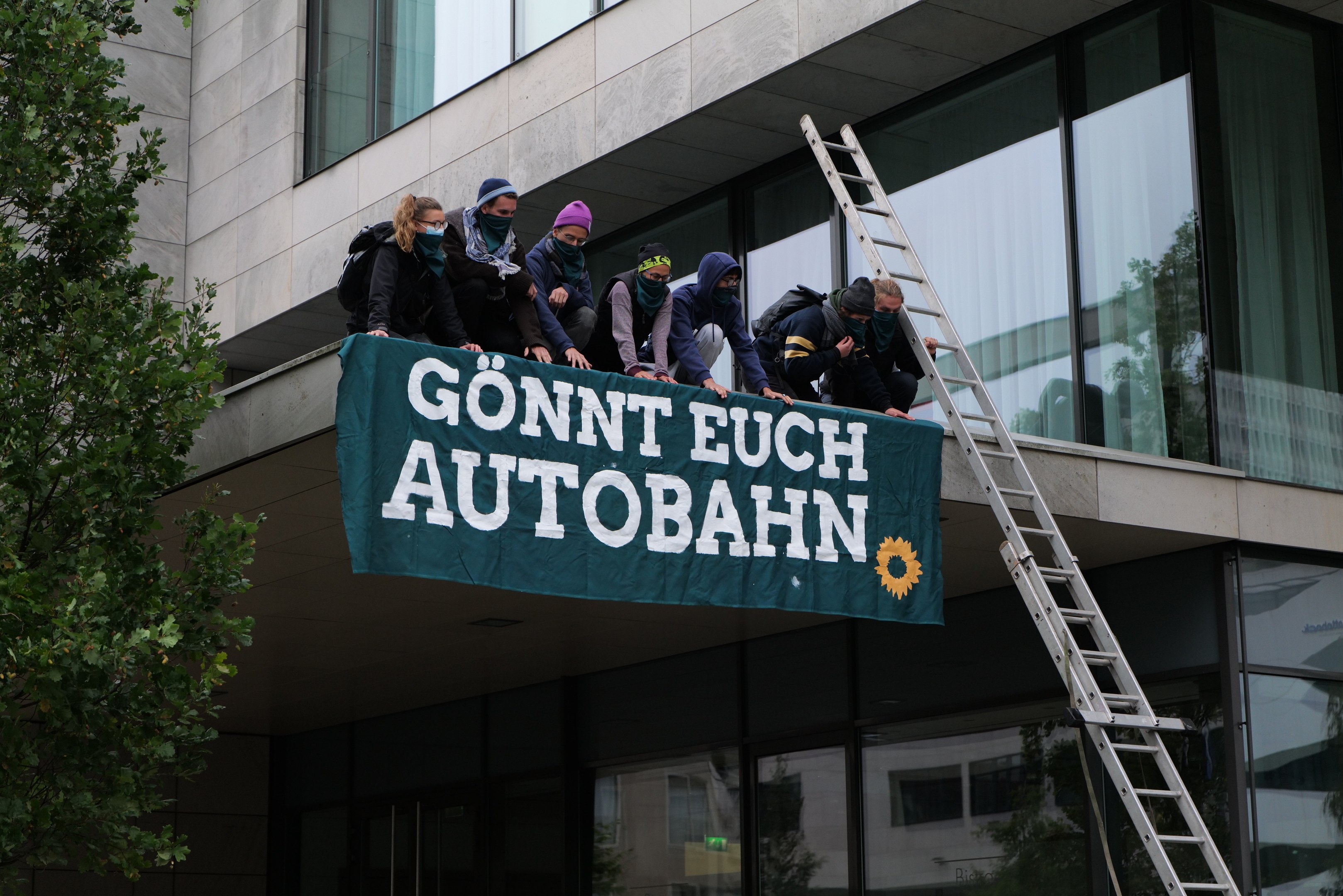 Menschen auf einem Gebäude mit einem Banner, einem Baum links und Glasfenstern im Hintergrund, die scheinbar für die deutsche Regierung protestieren.