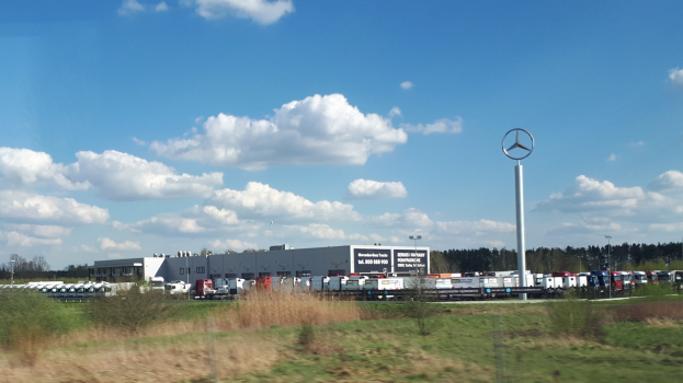 Mercedes-Benz-Fabrik in Deutschland mit Fahrzeugen auf der Straße, Grünfläche, Gebäuden und einem Logo-Pfahl unter einem bewölkten Himmel.