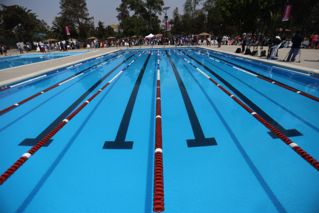 Gro├če Schwimmhalle mit Bahntrennern, umgeben von Menschen mit Taschen und Gegenst├Ąnden, mit Zelten, Bannern, Masten, einem Schild und einem bew├Âlktem Himmel im Hintergrund.