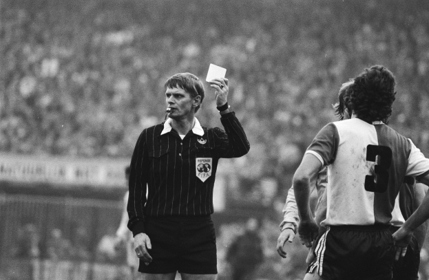 Schwarzes und weißes Bild eines Schiedsrichters mit roter Karte, mit einer Menge Menschen in Fußballuniformen hinter einem Zaun.