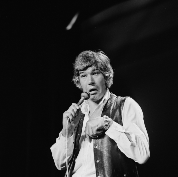 Ein Mann in weißem Hemd und schwarzer Weste singt leidenschaftlich in ein Mikrofon, seine Augen geschlossen und die Haare im klassischen Rocker-Look gestylt.