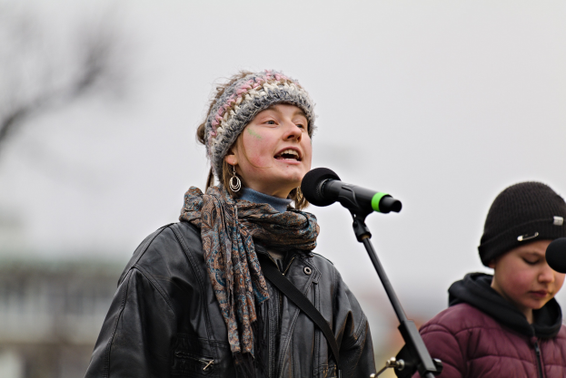 Eine Frau in einer schwarzen Jacke, einem Schal und einer Mütze singt in ein Mikrofon auf der Bühne, neben einer Person in einer marineblauen Jacke und einer schwarzen Mütze, während ein unscharfer Hintergrund die Szenerie bildet.