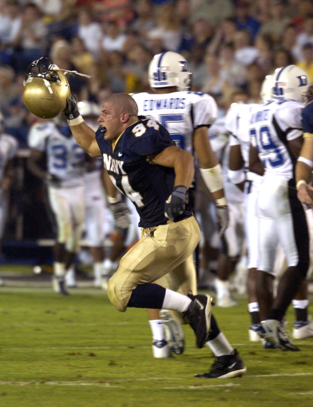 Ein Fußballspieler in dunkler Uniform rennt mit dem Ball, umgeben von Teamkollegen in Sportkleidung und Helmen auf dem Feld, während Zuschauer im Hintergrund auf Stühlen sitzen.