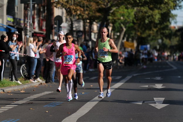 Gruppe von Menschen, die in einem Stadtmarathon laufen, Zuschauer links, unscharfer städtischer Hintergrund mit Bäumen, Gebäuden und einem Fahrrad.