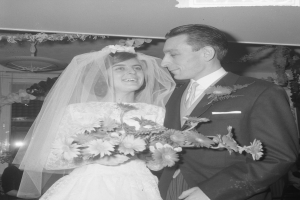 Ein Schwarz-Weiß-Foto eines Brautpaares, das zusammen steht, wobei die Braut einen Strauß hält und der Bräutigam einen Anzug trägt, sowie einige Menschen und dekorative Gegenstände im Hintergrund.