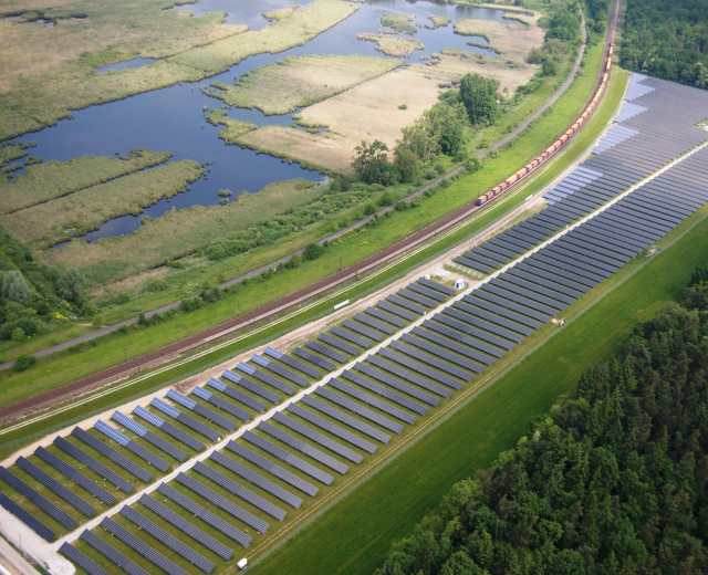Luftaufnahme einer Solaränlage mit Solarpanelen, umgeben von Bäumen, Gras, Pflanzen und Wasser, mit einem Zug auf einer näheren Eisenbahnschiene.