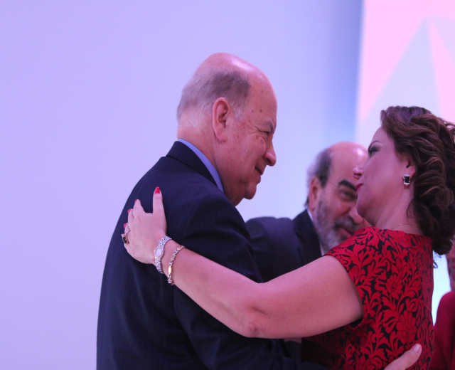 Zwei Menschen umarmen sich herzlich auf einer Bühne, umgeben von anderen, der Mann trägt ein Armband.