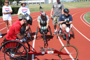 Eine Gruppe von Menschen im Rollstuhl auf einer Bahn umgeben von Gras, Pflanzen, Bäumen, Pfosten, Gebäuden und einem klaren blauen Himmel.