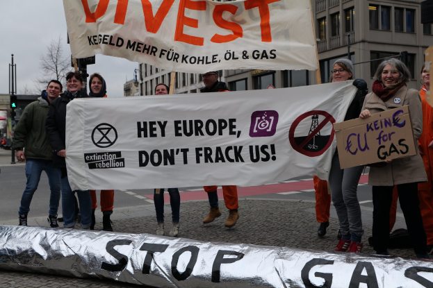 Eine Gruppe von Menschen auf einer Straße mit einem Banner, auf dem "Stop Gas" steht, protestiert gegen die Europäische Union. Im Hintergrund sind Gebäude, Bäume, Pfosten und Verkehrszeichen unter einem klaren blauen Himmel zu sehen.